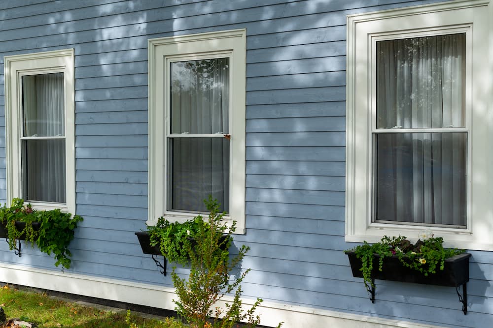The exterior wall of a building with three tall double hung windows and flower boxes. The powder blue horizontal narrow clapboard has shadows casts on it. The flower boxes have green plants hanging. Vinyl Siding, siding installation near me, siding replacement near me, siding companies chicago
