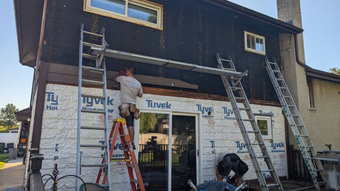 Workers are renovating the side of a house with ladders and Tyvek wrap.